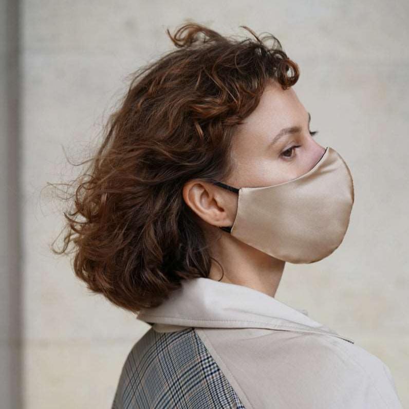 Beige Close-up of a woman wearing a French beige silk face mask with black adjustable straps, standing outdoors in a plaid coat.