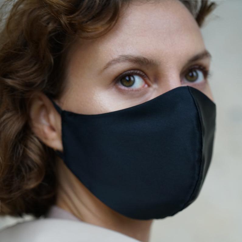 Side profile of a woman wearing a midnight black silk face mask, styled with loose curls and a beige trench coat