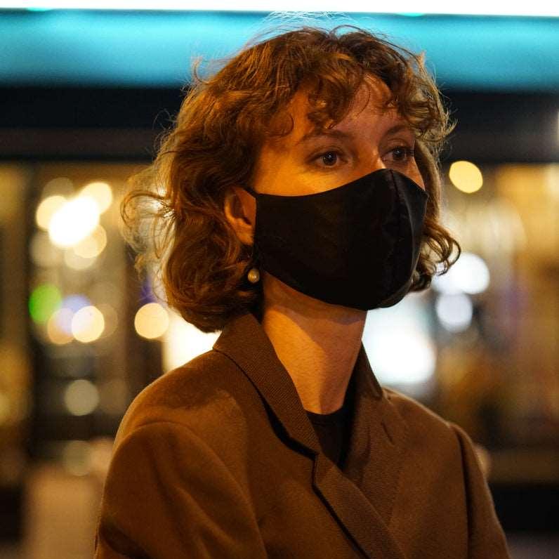 Stylish woman wearing a midnight black silk face mask in the evening, city lights reflecting in the background.