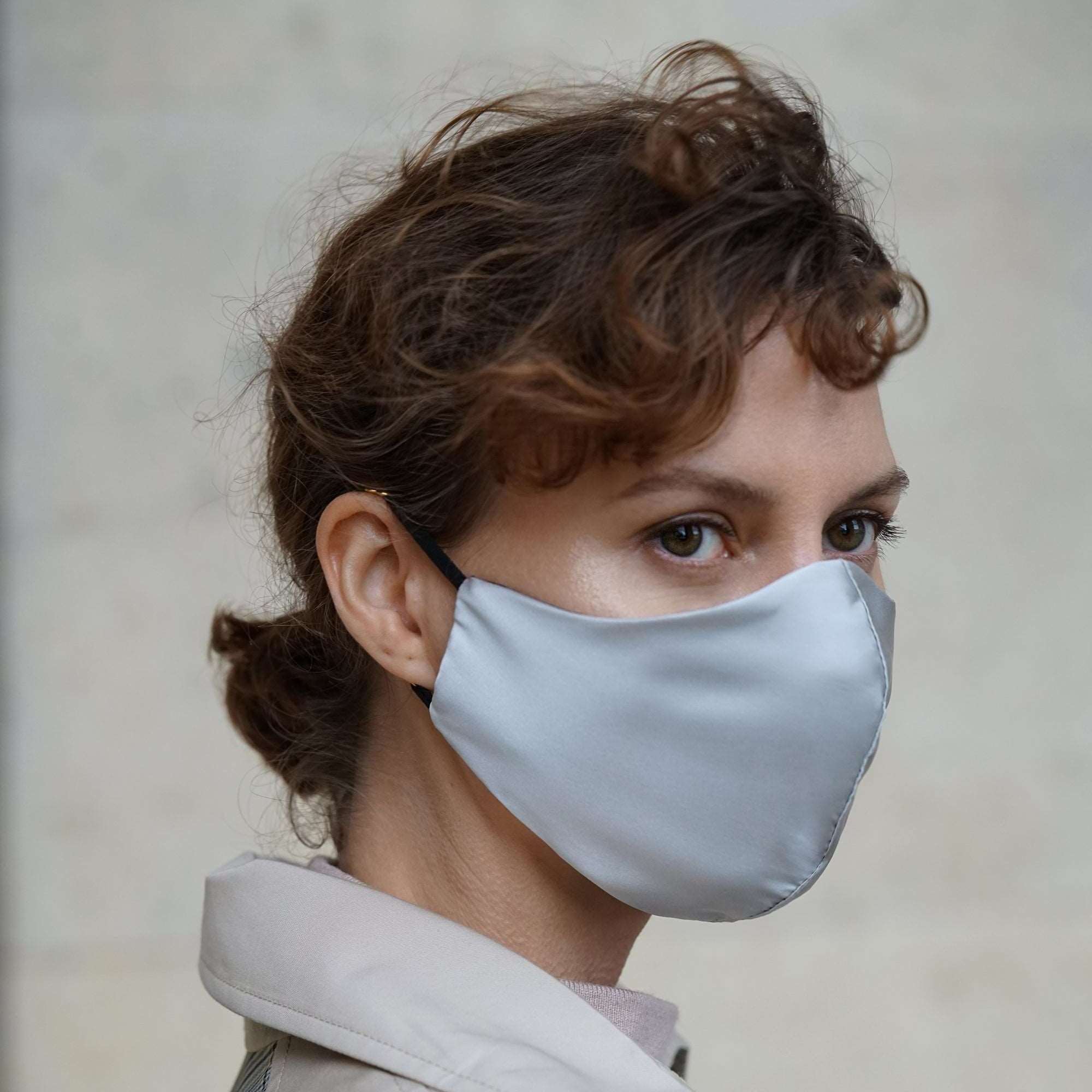 Grey Close-up of a woman wearing a silver grey silk face mask with black adjustable straps, paired with a beige trench coat.